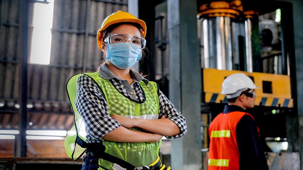 A woman in an industrial environment wearing a hard helmet and face mask